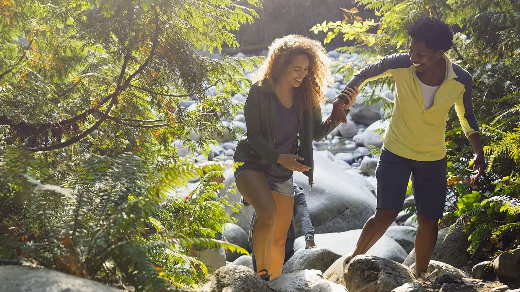 Drei Frauen beim Wandern, die zwei Frauen im Vordergrund helfen sich gegenseitig auf die Felsen zu klettern.